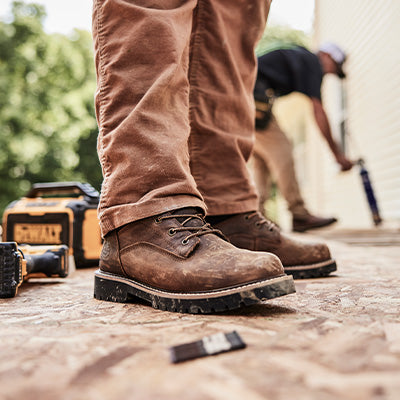 Close-up of brown work boots on a construction site with tools and another person in the background.
