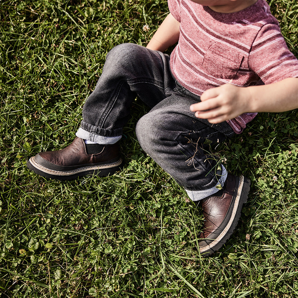 Child sitting on grass wearing brown boots and gray pants.