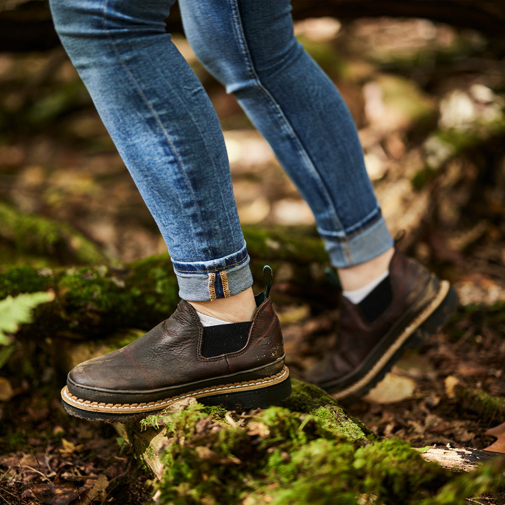 Person wearing blue jeans and brown shoes walking on a forest floor.