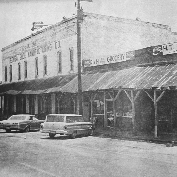 Vintage black and white photo of a street scene with storefronts and cars.