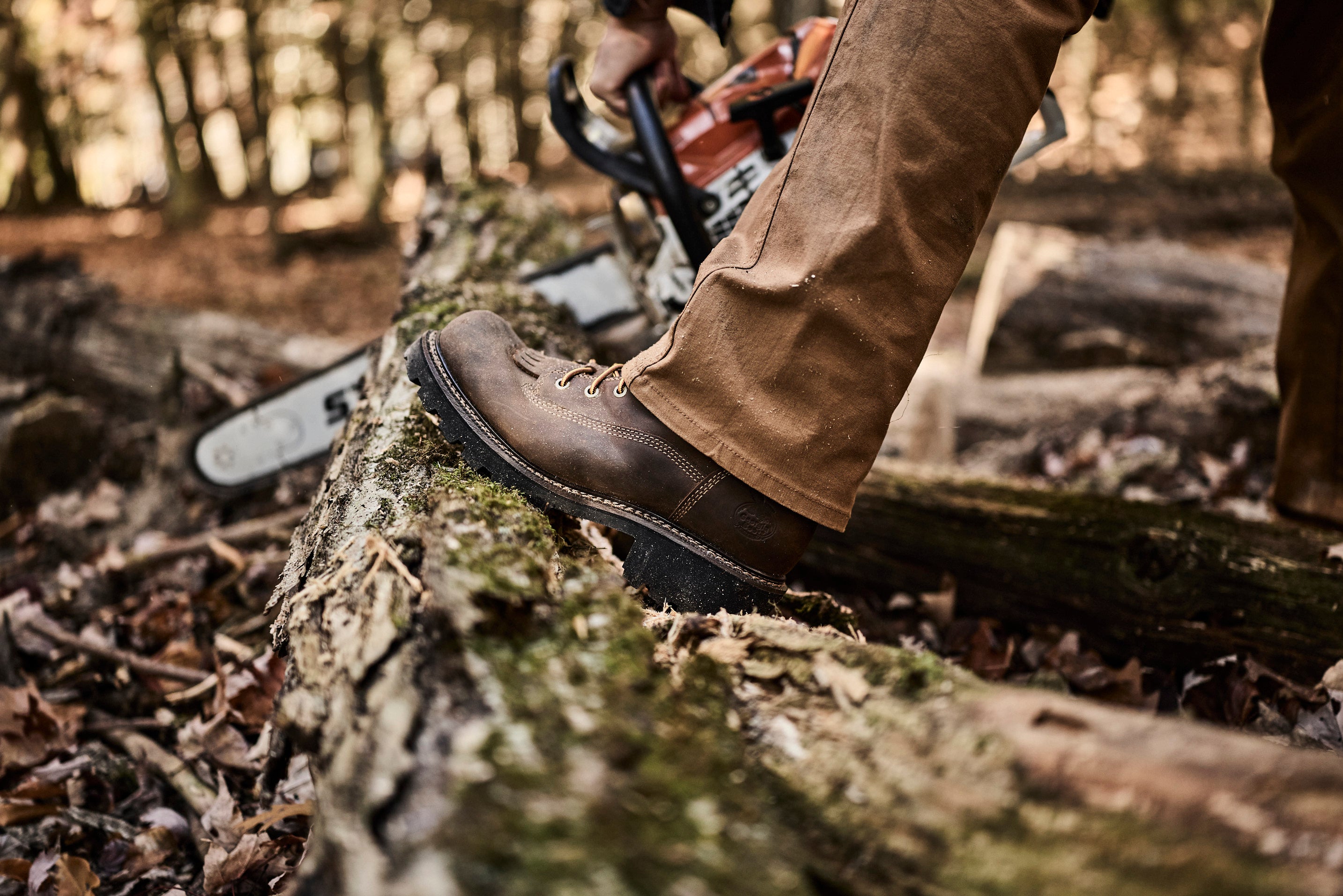 Person wearing brown boots and pants using a chainsaw on a log in a forest setting