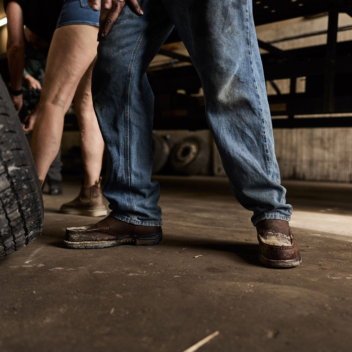 Close-up of a person wearing blue jeans and brown work boots next to a tire in an industrial setting.