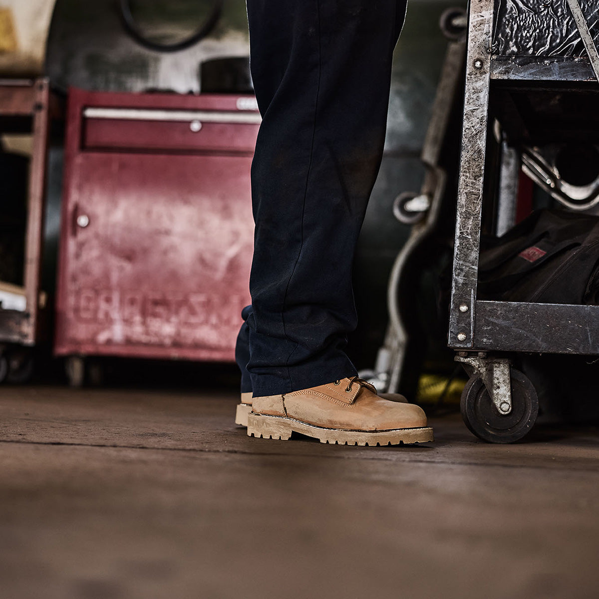 Person wearing tan work boots in a workshop setting with tools and equipment.
