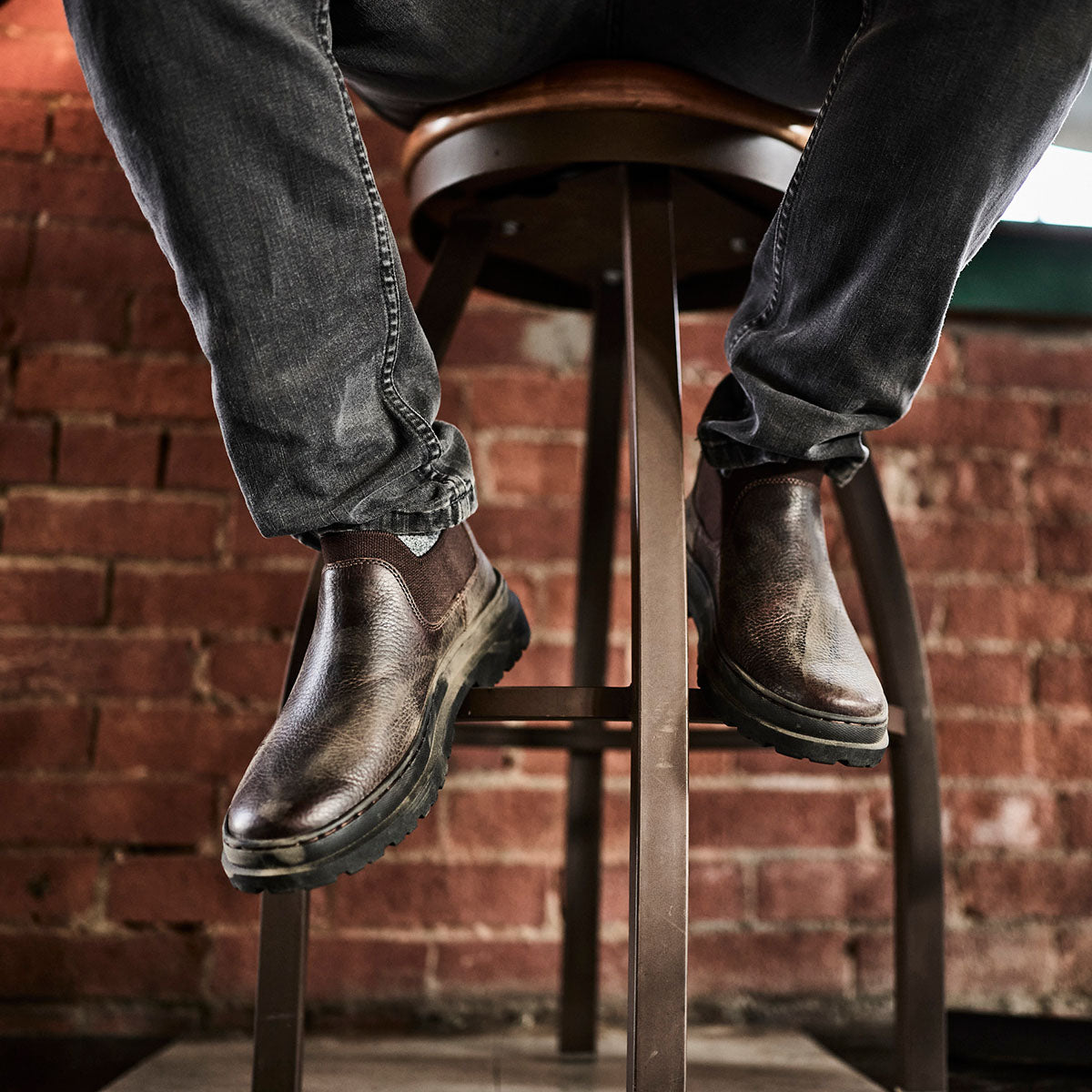 Person wearing brown leather boots and jeans sitting on a wooden stool against a brick wall.